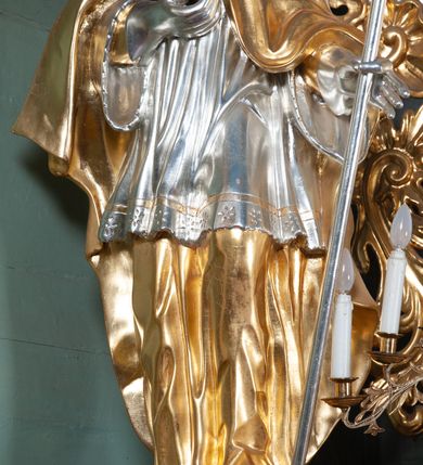 Zdjęcie nr 7: Ołtarz przyścienny, architektoniczny, na cokole, z bramkami. Nastawa jednoosiowa, jednokondygnacyjna ze zwieńczeniem. Mensa murowana, blat kamienny. Antepedium w kształcie leżącego prostokąta ujętego parami kolumn o gładkich trzonach i głowicach kompozytowych, podtrzymujących belkowanie z parami tryglifów. Belkowanie nad częścią środkową ucięte łukowato. W polu środkowym płaskorzeźbiona scena z przedstawieniem Świętej Rodziny. Postać św. Józefa półleżąca, zwrócona profilem w prawo, w stronę Matki Boskiej. Siedząca Maria skierowana w trzech czwartych w prawo, trzyma na kolanach poruszone Dzieciątko. Oś środkowa flankowana parą opilastrowanych filarów i kolumn o kapitelach kompozytowych, ustawionych przed licem ołtarza pod kątem. Podpory dźwigają pełne, wyłamane belkowanie oraz odpowiadający ich ułożeniu przerwany przyczółek. Zwieńczenie ujęte w spływy, zamknięte łukiem nadwieszonym. Cokoły kolumn dekorowane rocaille’em. W polu środkowym obraz we wnęce z przedstawieniem Matki Boskiej z Dzieciątkiem zasłaniany wizerunkiem św. Floriana w profilowanej ramie, w formie stojącego prostokąta zamkniętego dwoma, zwróconymi naprzeciwlegle stykającymi się spływami wolutowymi oraz rocaille’owym kartuszem z inskrypcją „POD TWOJĄ OBRONĘ”. Pierwsza kondygnacja ujęta uszakami utworzonymi z ornamentu rocaille’owego, ażurowej kratki regencyjnej i ornamentu roślinnego w formie kwiatów. Na bramkach ustawiono figury św. Wojciecha i św. Stanisława. Na przerwanym przyczółku spoczywa para adorujących aniołów. W zwieńczeniu profilowana rama powtarzająca jego formę z obrazem św. Antoniego Padewskiego zamknięta rocaille’em. Całość ujęta w spływy z ornamentu roślinnego. Struktura polichromowana na kremowo ze złoconą i srebrzoną ornamentyką oraz detalami architektonicznymi.