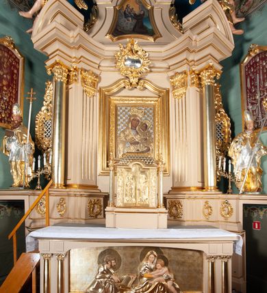 Zdjęcie nr 2: Tabernakulum w kształcie domku na podwójnym cokole, zwieńczonego dachem mansardowym, pokrytym dachówkami w formie łusek, z chwostami na krańcach kalenicy. Drzwiczki w kształcie stojącego prostokąta zakończonego łukiem nadwieszonym z płaskorzeźbionymi wizerunkami św. Piotra po lewej stronie i św. Pawła po prawej i aniołami na rewersie. Święci mają wydłużone proporcje i są ciasno owinięci płaszczami. Drzwiczki ujęte lizenami z dekoracją w formie festonów utworzonych z wstęgi regencyjnej, winnej latorośli i ornamentu roślinnego. 
Całość flankowana przez dwie wolnostojące kolumny o kanelowanych trzonach oraz kapitelach dekorowanych akantem i jajownikiem, dźwigających echinus z wazonem. 
Tabernakulum polichromowane na kremowo ze złoconymi drzwiczkami, dachem i kolumnami.