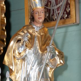 Zdjęcie nr 1: Rzeźba całopostaciowa, wydrążona. Postać zwrócona frontalnie, w kontrapoście, z lewą nogą wysuniętą do przodu, o esowato wygiętej sylwetce. W lewej ręce trzyma pastorał, prawą unosi w geście błogosławieństwa. Głowa delikatnie przechylona na prawe ramię. Włosy w postaci długich pukli spływających na plecy. Twarz pełna, o wyraźnych rysach. Na głowie infuła udekorowana rozetą. Święty w stroju biskupim: albie, rokiecie i rękawiczkach; na nogach buty. Szaty ekspresyjnie poruszone, odzwierciedlające układ sylwetki, w formie drobnych, głęboko rzeźbionych fałd. 
Partie ciała polichromowane naturalistycznie, zaś szaty, atrybuty złocone i srebrzone.