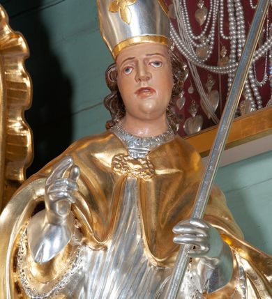 Zdjęcie nr 2: Rzeźba całopostaciowa, wydrążona. Postać zwrócona frontalnie, w kontrapoście, z lewą nogą wysuniętą do przodu, o esowato wygiętej sylwetce. W lewej ręce trzyma pastorał, prawą unosi w geście błogosławieństwa. Głowa delikatnie przechylona na prawe ramię. Włosy w postaci długich pukli spływających na plecy. Twarz pełna, o wyraźnych rysach. Na głowie infuła udekorowana rozetą. Święty w stroju biskupim: albie, rokiecie i rękawiczkach; na nogach buty. Szaty ekspresyjnie poruszone, odzwierciedlające układ sylwetki, w formie drobnych, głęboko rzeźbionych fałd. 
Partie ciała polichromowane naturalistycznie, zaś szaty, atrybuty złocone i srebrzone.