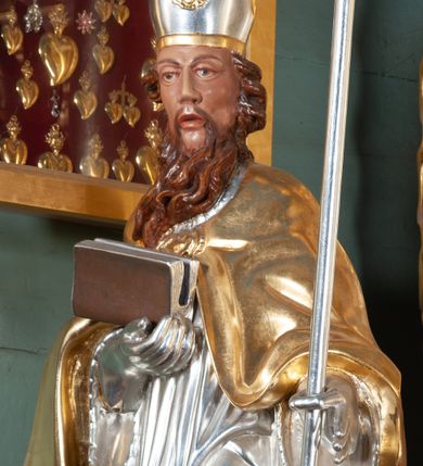 Zdjęcie nr 1: Rzeźba całopostaciowa, drążona. Postać ustawiona frontalnie, w kontrapoście, z lewą nogą wysuniętą do przodu. W lewej ręce trzyma krzyż arcybiskupi, prawą ręką przytrzymuje przy piersi księgę. Głowa zwrócona delikatnie w prawo, twarz szczupła o wyraźnych rysach, okolona długą brodą w postaci pukli spływających w stronę prawego ramienia. Święty ma brązowe oczy, długi nos i rozchylone usta. Jest ubrany w albę, komżę dekorowaną u spodu ornamentem roślinnym oraz kapę zapiętą na piersi fibulą, na głowie ma infułę dekorowaną rozetą, na rękach rękawiczki, spod szat widać czubki butów. Szaty miękko kształtowane o drobnych fałdach, niekiedy ściśle przylegających do ciała, odzwierciedlające ruch postaci. Partie ciała polichromowane naturalistyczne, szaty i atrybuty złocone oraz srebrzone.