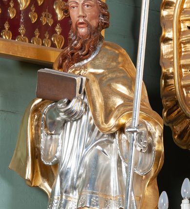 Zdjęcie nr 2: Rzeźba całopostaciowa, drążona. Postać ustawiona frontalnie, w kontrapoście, z lewą nogą wysuniętą do przodu. W lewej ręce trzyma krzyż arcybiskupi, prawą ręką przytrzymuje przy piersi księgę. Głowa zwrócona delikatnie w prawo, twarz szczupła o wyraźnych rysach, okolona długą brodą w postaci pukli spływających w stronę prawego ramienia. Święty ma brązowe oczy, długi nos i rozchylone usta. Jest ubrany w albę, komżę dekorowaną u spodu ornamentem roślinnym oraz kapę zapiętą na piersi fibulą, na głowie ma infułę dekorowaną rozetą, na rękach rękawiczki, spod szat widać czubki butów. Szaty miękko kształtowane o drobnych fałdach, niekiedy ściśle przylegających do ciała, odzwierciedlające ruch postaci. Partie ciała polichromowane naturalistyczne, szaty i atrybuty złocone oraz srebrzone.