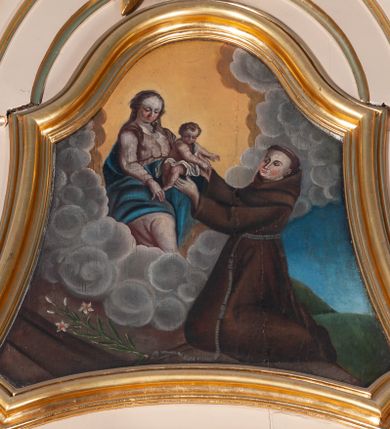 Zdjęcie nr 1: Pole obrazowe w kształcie wklęsło-wypukłym. W centrum scena, w której klęczący św. Antoni odbiera Dzieciątko Jezus od ukazującej mu się na obłokach Marii. Święty ma okrągłą twarz, wąski nos i rumieńce, na głowie wyciętą tonsurę, jest ubrany w brązowy habit franciszkański. Matka Boska siedzi, prawą rękę ma położoną na udzie, patrzy na rozgrywającą się scenę. Ma okrągłą twarz i długie włosy spływające na ramiona i plecy. Jest ubrana w różową suknię, niebieski płaszcz oraz zarzucony na głowę, przezroczysty welon. Dzieciątko wyciąga ręce w stronę św. Antoniego, jest przepasane białą tkaniną. U dołu sceny po lewej stronie leży kwiat lilii. Tło zróżnicowane: za Matką Boską żółte, za św. Antonim niebieskie, między nimi kłębią się szare obłoki, poniżej nich podest; silny światłocień. 
