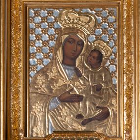 Zdjęcie nr 1: Obraz w kształcie stojącego prostokąta z przedstawieniem Marii ujętej w półfigurze, zwróconej nieznacznie w lewo, z Dzieciątkiem Jezus na lewej ręce. Maria lewą ręką wskazuje na Jezusa, prawą podtrzymuje jego nogi, patrzy na widza. Ma owalną twarz, brązowe oczy, wąski nos i małe, wydatne usta. Dzieciątko patrzy na Matkę, prawą ręką czyni gest błogosławieństwa, lewą przytrzymuje zamkniętą księgę. Ma owalną twarz, prosty nos i wąskie usta oraz krótkie, brązowe włosy. Karnacja obu postaci jest ciemna, światłocień delikatny. Resztę obrazu przykrywają srebrne, złocone sukienki oraz tło. Maria ubrana jest w srebrną suknię oraz złoty, przykrywający głowę płaszcz dekorowany rozwiniętymi kwiatami, spięty pod szyją. Dzieciątko ma na sobie długą, złotą, przewiązaną w pasie sukienkę. Oboje mają na głowach zamknięte korony. Tło dekorowane kratką, na przecięciu listewek rozetki. Obraz ujęty złotą ramą zdobioną wicią roślinną i rocaille’em. 