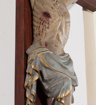 Zdjęcie nr 5: Pełnoplastyczna, drążona od tyłu, polichromowana figura Chrystusa przybita do prostego krzyża trzema gwoździami. Sylwetka zmarłego Chrystusa smukła z wyraźnie zaznaczoną muskulaturą i głębokim wcięciem w talii i widocznym układem kostnym klatki piersiowej. Ramiona uniesione wysoko tworzą łuk, dłonie o wyprostowanych palcach. Nogi znacznie ugięte w kolanach, założone prawa na lewą. Głowa pochylona, lekko zwrócona w prawą stronę. Na głowie Chrystus ma założoną pozłacaną koronę cierniową. Twarz podłużna z zapadniętymi policzkami, oczy zamknięte z opadłymi kącikami i ściągniętymi brwiami, nos długi o prostym profilu, usta małe i zamknięte. Włosy długie, opadające na kark, broda krótka i kręcona. Perizonium długie, sięgające połowy łydki, po bokach opadające swobodnie kaskadowymi fałdami. Polichromowane na kolor szary, na brzegu złocone. Nad głową titulus w formie poziomej banderoli z napisem „I.N.R.I.”. W odsłoniętych partiach ciała figura polichromowana naturalistycznie o ciemnej karnacji z zaznaczonymi śladami męki. Krzyż szeroki, prosty, pokryty bejcą. 
