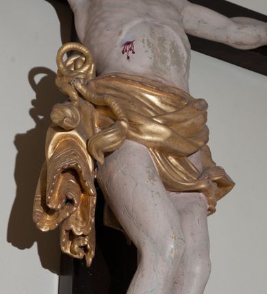 Zdjęcie nr 4: Rzeźba Chrystusa przybita do krzyża czterema gwoździami, ukazana w typie Cristo morte. Ciało o szeroko rozstawionych ramionach, sylwetka szczupła, choć muskularna. Chrystus ma głowę przechyloną na prawe ramię. Twarz szczupła, z wąskim i długim nosem, zamkniętymi oczami, na wpół otwartymi ustami, okolona krótką brodą. Włosy długie, ciemnobrązowe spływające na prawe ramię, na głowie zielona korona cierniowa. Perizonium złocone, zdobione na brzegu koronką, swobodnie oplatające biodra, przewiązane sznurem na prawym boku, z jednym końcem dłuższym, zwisającym wzdłuż prawego uda. Z boku, dłoni, stóp i spod korony cierniowej spływają strużki krwi. Polichromia ciała naturalistyczna. Krzyż gładki, prosty, pomalowany na ciemnobrązowy kolor; nad głową Chrystusa titulus z napisem „INRI” w języku łacińskim, greckim i hebrajskim. U podstawy pionowej belki krzyża złocona i marmoryzowana, wolutowo wygięta konsolka. 
