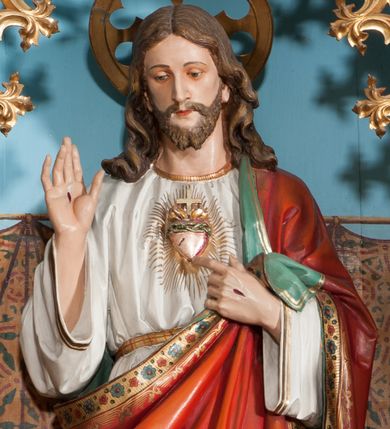 Zdjęcie nr 1: Rzeźba pełnoplastyczna, ustawiona na niskim prostopadłościennym cokole. Chrystus ukazany w całej postaci, stojący w delikatnym kontrapoście, prawą ręką czyni gest błogosławieństwa, lewą wskazuje na gorejące na piersi serce. Głowa delikatnie skłoniona w dół, twarz podłużna z długim i wąskim nosem, okolona krótką brodą, rozdzieloną na dwa pukle. Włosy długie, ciemnobrązowe, z przedziałkiem pośrodku, opadające na plecy bujnymi falami. Chrystus ubrany jest w długą białą suknię, obszytą na brzegach złotą lamówką, przewiązaną w talii złotym paskiem oraz czerwony płaszcz o oliwkowozielonej podszewce, bogato zdobiony stylizowanymi kwiatami, założony na lewy bark, drugi koniec płaszcza przytrzymuje lewą ręką, na stopach sandały. Płaszcz dodatkowo dekorowany jest na brzegu ozdobną lamówką złożoną z rzędu wielopłatkowych kwiatków. Na dłoniach Chrystus ma widoczne ślady męki, a na piersi gorejące serce, oplecione koroną cierniową, z zatkniętym u góry krzyżykiem, ukazane na tle promienistej glorii. Polichromia w odsłoniętych partiach ciała naturalistyczna, nad głową Chrystusa okrągły nimb, z wyciętym ażurowo w polu krzyżem.


