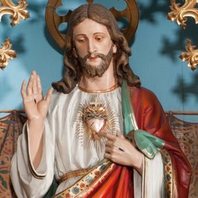 Zdjęcie nr 1: Rzeźba pełnoplastyczna, ustawiona na niskim prostopadłościennym cokole. Chrystus ukazany w całej postaci, stojący w delikatnym kontrapoście, prawą ręką czyni gest błogosławieństwa, lewą wskazuje na gorejące na piersi serce. Głowa delikatnie skłoniona w dół, twarz podłużna z długim i wąskim nosem, okolona krótką brodą, rozdzieloną na dwa pukle. Włosy długie, ciemnobrązowe, z przedziałkiem pośrodku, opadające na plecy bujnymi falami. Chrystus ubrany jest w długą białą suknię, obszytą na brzegach złotą lamówką, przewiązaną w talii złotym paskiem oraz czerwony płaszcz o oliwkowozielonej podszewce, bogato zdobiony stylizowanymi kwiatami, założony na lewy bark, drugi koniec płaszcza przytrzymuje lewą ręką, na stopach sandały. Płaszcz dodatkowo dekorowany jest na brzegu ozdobną lamówką złożoną z rzędu wielopłatkowych kwiatków. Na dłoniach Chrystus ma widoczne ślady męki, a na piersi gorejące serce, oplecione koroną cierniową, z zatkniętym u góry krzyżykiem, ukazane na tle promienistej glorii. Polichromia w odsłoniętych partiach ciała naturalistyczna, nad głową Chrystusa okrągły nimb, z wyciętym ażurowo w polu krzyżem.


