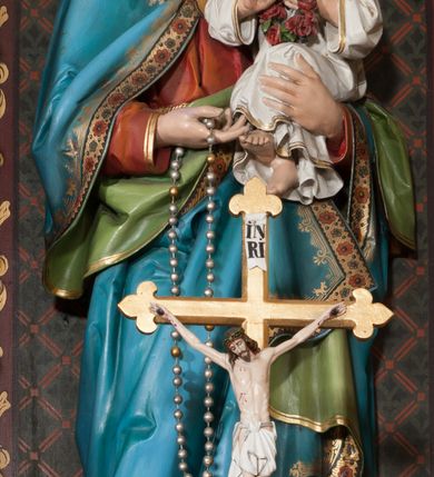 Zdjęcie nr 1: Rzeźba pełnoplastyczna, ustawiona na niskim cokole, w delikatnym kontrapoście. Matka Boska trzyma na lewym ręku Dzieciątko Jezus, w prawej różaniec, a głowę delikatnie przechyla na lewą stronę. Ma owalną twarz, przymknięte oczy ze wzrokiem skierowanym w dół, długi i prosty nos oraz zarumienione policzki. Długie, brązowe włosy opadają grubymi lokami na plecy i ramiona. Maria ubrana jest w długą, czerwoną suknię z długimi rękawami oraz błękitny płaszcz z zieloną podszewką, spięty na piersiach złotą broszą, na głowie ma biały welon i koronę w typie otwartym. Szaty, a zwłaszcza płaszcz obwiedzione są złoconą lamówką i dekorowane ornamentalnym pasem. Dzieciątko ukazane jest w pozycji siedzącej, zwrócone trzy czwarte w prawo, w dłoniach trzyma wianek z czerwonych róż. Ma idealizowane rysy twarzy oraz krótkie, jasne i kędzierzawe włosy. Ubrane jest w długą, białą sukienkę z długimi rękawami. Polichromia w odsłoniętych partiach ciała naturalistyczna. 
