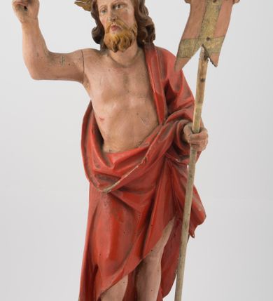 Zdjęcie nr 1: Rzeźba Chrystusa Zmartwychwstałego, ścięta z tyłu, ustawiona na niskim, zdwojonym, prostopadłościennym cokole. Chrystus ukazany  frontalnie, w kontrapoście, delikatnie przechylony w prawą stronę, w lewej ręce trzyma chorągiew zakończoną krzyżem, a prawą błogosławi. Twarz pociągła, długi i wąski nos, broda jasna, średniej długości. Włosy długie, brązowe, z przedziałkiem na środku głowy, opadają na ramiona w postaci loków, zasłaniając uszy. Chrystus ubrany jest w czerwony płaszcz, przewieszony przez lewe ramię, odsłaniający nagi tors.  Nimb krzyżowy w formie trzech wiązek promieni o nierównej długości. Polichromia naturalistyczna, nimb złocony.


