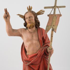 Zdjęcie nr 1: Rzeźba Chrystusa Zmartwychwstałego, ścięta z tyłu, ustawiona na niskim, zdwojonym, prostopadłościennym cokole. Chrystus ukazany  frontalnie, w kontrapoście, delikatnie przechylony w prawą stronę, w lewej ręce trzyma chorągiew zakończoną krzyżem, a prawą błogosławi. Twarz pociągła, długi i wąski nos, broda jasna, średniej długości. Włosy długie, brązowe, z przedziałkiem na środku głowy, opadają na ramiona w postaci loków, zasłaniając uszy. Chrystus ubrany jest w czerwony płaszcz, przewieszony przez lewe ramię, odsłaniający nagi tors.  Nimb krzyżowy w formie trzech wiązek promieni o nierównej długości. Polichromia naturalistyczna, nimb złocony.


