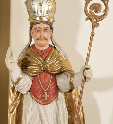 Zdjęcie nr 1: Rzeźba ścięta z tyłu, przedstawia św. Stanisława. Święty ukazany w postawie stojącej na niskim cokole, frontalnie, w statycznej pozie, z rękami wyciągniętymi do przodu i odwiedzionymi delikatnie na boki: prawą wykonuje gest błogosławieństwa, w lewej trzyma pastorał. Ma szeroką twarz, z długim i prostym nosem, wzrokiem skierowanym na wprost oraz wąsy. Włosy krótkie, kędzierzawe, ciemnobrązowe. Święty jest ubrany jest w czerwoną sutannę, białą rokietę zdobioną na brzegach złoceniem, czerwony mucet, złotą kapę, spiętą na piersiach broszą, białe rękawice, jasnobrązowe buty oraz srebrzoną infułę, zdobioną złoconymi kaboszonami, rautami, motywami w kształcie litery „S” oraz czteropłatkowymi kwiatami. Na piersi ma zawieszony złocony krzyż pektoralny. Obok jego prawej nogi znajduje się mała klęcząca postać Piotrowina ze złożonymi rękami, ubranego w białą, długą suknię i zielony płaszcz. Polichromia w odsłoniętych partiach ciała jest naturalistyczna. Cokół jasnozielony, z przodu czarno malowany napis: „ŚW(IĘTY) STANISŁAW.” 




