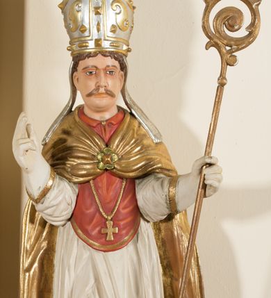 Zdjęcie nr 1: Rzeźba ścięta z tyłu, przedstawia św. Stanisława. Święty ukazany w postawie stojącej na niskim cokole, frontalnie, w statycznej pozie, z rękami wyciągniętymi do przodu i odwiedzionymi delikatnie na boki: prawą wykonuje gest błogosławieństwa, w lewej trzyma pastorał. Ma szeroką twarz, z długim i prostym nosem, wzrokiem skierowanym na wprost oraz wąsy. Włosy krótkie, kędzierzawe, ciemnobrązowe. Święty jest ubrany jest w czerwoną sutannę, białą rokietę zdobioną na brzegach złoceniem, czerwony mucet, złotą kapę, spiętą na piersiach broszą, białe rękawice, jasnobrązowe buty oraz srebrzoną infułę, zdobioną złoconymi kaboszonami, rautami, motywami w kształcie litery „S” oraz czteropłatkowymi kwiatami. Na piersi ma zawieszony złocony krzyż pektoralny. Obok jego prawej nogi znajduje się mała klęcząca postać Piotrowina ze złożonymi rękami, ubranego w białą, długą suknię i zielony płaszcz. Polichromia w odsłoniętych partiach ciała jest naturalistyczna. Cokół jasnozielony, z przodu czarno malowany napis: „ŚW(IĘTY) STANISŁAW.” 




