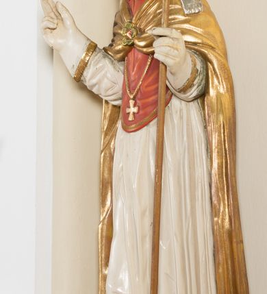 Zdjęcie nr 3: Rzeźba ścięta z tyłu, przedstawia św. Stanisława. Święty ukazany w postawie stojącej na niskim cokole, frontalnie, w statycznej pozie, z rękami wyciągniętymi do przodu i odwiedzionymi delikatnie na boki: prawą wykonuje gest błogosławieństwa, w lewej trzyma pastorał. Ma szeroką twarz, z długim i prostym nosem, wzrokiem skierowanym na wprost oraz wąsy. Włosy krótkie, kędzierzawe, ciemnobrązowe. Święty jest ubrany jest w czerwoną sutannę, białą rokietę zdobioną na brzegach złoceniem, czerwony mucet, złotą kapę, spiętą na piersiach broszą, białe rękawice, jasnobrązowe buty oraz srebrzoną infułę, zdobioną złoconymi kaboszonami, rautami, motywami w kształcie litery „S” oraz czteropłatkowymi kwiatami. Na piersi ma zawieszony złocony krzyż pektoralny. Obok jego prawej nogi znajduje się mała klęcząca postać Piotrowina ze złożonymi rękami, ubranego w białą, długą suknię i zielony płaszcz. Polichromia w odsłoniętych partiach ciała jest naturalistyczna. Cokół jasnozielony, z przodu czarno malowany napis: „ŚW(IĘTY) STANISŁAW.” 




