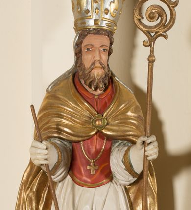 Zdjęcie nr 1: Rzeźba ścięta z tyłu, przedstawia św. Wojciecha. Święty ukazany w postawie stojącej na niskim cokole, frontalnie, w statycznej pozie, z rękami wyciągniętymi do przodu, w których trzyma: w prawej wiosło, w lewej pastorał. Ma podłużną twarz, z długim i prostym nosem, wzrokiem skierowanym na wprost, okoloną średniej długości brodą. Włosy krótkie, kędzierzawe, zasłaniające uszy. Święty jest ubrany w czerwoną albę, białą rokietę zdobioną na brzegach złoceniem, czerwony mucet, złotą kapę, spiętą na piersiach broszą, białe rękawice, jasnobrązowe buty oraz srebrzoną infułę, zdobioną złoconymi kaboszonami, rautami, motywami w kształcie litery „S” oraz czteropłatkowymi kwiatami. Na piersi ma zawieszony złocony krzyż pektoralny. Polichromia w odsłoniętych partiach ciała naturalistyczna. Cokół jasnozielony, z przodu czarno malowany napis: „ŚW(IĘTY) WOJCIECH.”
