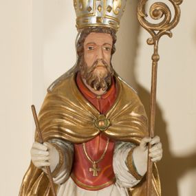 Zdjęcie nr 1: Rzeźba ścięta z tyłu, przedstawia św. Wojciecha. Święty ukazany w postawie stojącej na niskim cokole, frontalnie, w statycznej pozie, z rękami wyciągniętymi do przodu, w których trzyma: w prawej wiosło, w lewej pastorał. Ma podłużną twarz, z długim i prostym nosem, wzrokiem skierowanym na wprost, okoloną średniej długości brodą. Włosy krótkie, kędzierzawe, zasłaniające uszy. Święty jest ubrany w czerwoną albę, białą rokietę zdobioną na brzegach złoceniem, czerwony mucet, złotą kapę, spiętą na piersiach broszą, białe rękawice, jasnobrązowe buty oraz srebrzoną infułę, zdobioną złoconymi kaboszonami, rautami, motywami w kształcie litery „S” oraz czteropłatkowymi kwiatami. Na piersi ma zawieszony złocony krzyż pektoralny. Polichromia w odsłoniętych partiach ciała naturalistyczna. Cokół jasnozielony, z przodu czarno malowany napis: „ŚW(IĘTY) WOJCIECH.”
