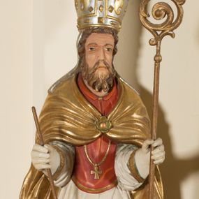 Zdjęcie nr 1: Rzeźba ścięta z tyłu, przedstawia św. Wojciecha. Święty ukazany w postawie stojącej na niskim cokole, frontalnie, w statycznej pozie, z rękami wyciągniętymi do przodu, w których trzyma: w prawej wiosło, w lewej pastorał. Ma podłużną twarz, z długim i prostym nosem, wzrokiem skierowanym na wprost, okoloną średniej długości brodą. Włosy krótkie, kędzierzawe, zasłaniające uszy. Święty jest ubrany w czerwoną albę, białą rokietę zdobioną na brzegach złoceniem, czerwony mucet, złotą kapę, spiętą na piersiach broszą, białe rękawice, jasnobrązowe buty oraz srebrzoną infułę, zdobioną złoconymi kaboszonami, rautami, motywami w kształcie litery „S” oraz czteropłatkowymi kwiatami. Na piersi ma zawieszony złocony krzyż pektoralny. Polichromia w odsłoniętych partiach ciała naturalistyczna. Cokół jasnozielony, z przodu czarno malowany napis: „ŚW(IĘTY) WOJCIECH.”
