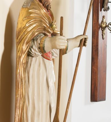 Zdjęcie nr 3: Rzeźba ścięta z tyłu, przedstawia św. Wojciecha. Święty ukazany w postawie stojącej na niskim cokole, frontalnie, w statycznej pozie, z rękami wyciągniętymi do przodu, w których trzyma: w prawej wiosło, w lewej pastorał. Ma podłużną twarz, z długim i prostym nosem, wzrokiem skierowanym na wprost, okoloną średniej długości brodą. Włosy krótkie, kędzierzawe, zasłaniające uszy. Święty jest ubrany w czerwoną albę, białą rokietę zdobioną na brzegach złoceniem, czerwony mucet, złotą kapę, spiętą na piersiach broszą, białe rękawice, jasnobrązowe buty oraz srebrzoną infułę, zdobioną złoconymi kaboszonami, rautami, motywami w kształcie litery „S” oraz czteropłatkowymi kwiatami. Na piersi ma zawieszony złocony krzyż pektoralny. Polichromia w odsłoniętych partiach ciała naturalistyczna. Cokół jasnozielony, z przodu czarno malowany napis: „ŚW(IĘTY) WOJCIECH.”
