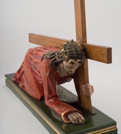 Zdjęcie nr 3: Pełnoplastyczna, wolnostojąca rzeźba Chrystusa upadającego pod krzyżem ustawiona na niskim, prostopadłościennym postumencie. Chrystus przedstawiony jest w pozycji półklęczącej, z prawą nogą ugiętą w kolanie, wysuniętą do przodu, lewą wyprostowaną do tyłu, z ciałem podpartym na ugiętym w łokciu prawym ramieniu. Lewą ręką podtrzymuje krzyż. Twarz pociągła, okolona ciemnym zarostem oraz długimi opadającymi na plecy i ramiona włosami. Chrystus jest ubrany w długą, czerwoną szatę przepasaną złotym sznurem. Na głowie złocona cierniowa korona. Figura w pełni polichromowana.
