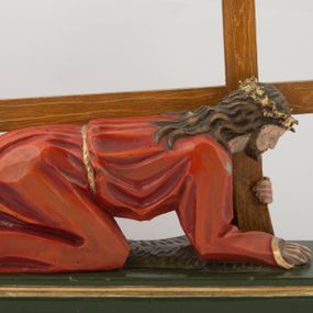 Zdjęcie nr 1: Pełnoplastyczna, wolnostojąca rzeźba Chrystusa upadającego pod krzyżem ustawiona na niskim, prostopadłościennym postumencie. Chrystus przedstawiony jest w pozycji półklęczącej, z prawą nogą ugiętą w kolanie, wysuniętą do przodu, lewą wyprostowaną do tyłu, z ciałem podpartym na ugiętym w łokciu prawym ramieniu. Lewą ręką podtrzymuje krzyż. Twarz pociągła, okolona ciemnym zarostem oraz długimi opadającymi na plecy i ramiona włosami. Chrystus jest ubrany w długą, czerwoną szatę przepasaną złotym sznurem. Na głowie złocona cierniowa korona. Figura w pełni polichromowana.
