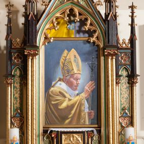 Zdjęcie nr 1: Niewielki ołtarz przyścienny, architektoniczny o formach neogotyckich, trójosiowy, jednokondygnacyjny, ustawiony na niskim cokole. Tabernakulum w formie prostopadłościennej skrzynki, ujęte złoconymi kolumienkami wspierającymi gzyms. Drzwiczki w kształcie stojącego prostokąta z płaskorzeźbioną dekoracją mi.n. z motywem czwórliścia. Nastawa ujęta w osi głównej zdwojonymi smukłymi, kolumienkami o złoconych trzonach, dźwigających ostrołukową arkadę i pinakle na osi kolumn. W polu obraz w kształcie stojącego prostokąta z przedstawieniem św. Jana Pawła II, poniżej wąskie płyciny z malowaną roślinną dekoracją na zielonym tle. Osie boczne nieco niższe, znacznie węższe z ostrołukowymi płycinami w polach ujętymi kolumienkami. W płycinach malowana dekoracja roślinna; w zwieńczeniach pinakle z czołgankami i kwiatonami. W zwieńczeniu wimperga wypełniona maswerkiem z motywem wieloliścia oraz rybiego pęcherza. U szczytu pinakiel z kwiatonem. Struktura malowana na kolor ciemnobrązowy, profilowania złocone i malowane na kolory niebieski oraz zielony, ornamenty złocone, płyciny malowane na czerwono i zielono.