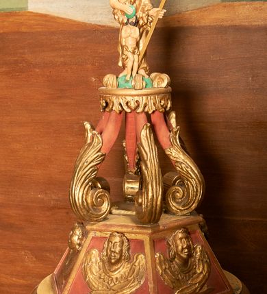 Zdjęcie nr 2: Chrzcielnica ustawiona podstawie z uskokiem. Trzon tworzy gruszkowaty nodus z pierścieniem. Czara puklowana, o wywiniętym wylewie. Pokrywa na planie koła, przechodząca w sześciobok ze ścianami dekorowanymi uskrzydlonymi główkami anielskimi. Na niej ustawiono sześć pałąków zdobionych wolutowo skręconymi liśćmi akantu, dźwigających podstawę dla grupy rzeźbiarskiej z przedstawieniem chrztu Chrystusa. Frontalnie ustawiony Jan Chrzciciel jest ubrany w melotę, w lewej ręce trzyma krzyż, w prawej przechyloną misę. Przed nim nieproporcjonalnie mała postać Chrystusa w kontrapoście, z dłońmi złożonymi w geście modlitwy.