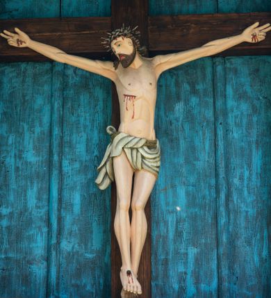 Zdjęcie nr 1: Krucyfiks drewniany w kapliczkowej oprawie umieszczony na absydzie, ponad sobotami. Chrystus przybity trzema gwoździami. Głowa opada na prawe ramię, oczy otwarte, nos długi, usta o kącikach skierowanych w dół. Twarz okala brązowy zarost. Na głowie ciemna korona cierniowa, włosy brązowe spływające na plecy i prawe ramię. Ciało umięśnione, nogi lekko ugięte. Z przebitego boku, spod korony oraz z miejsc przebicia gwoździami spływają strużki krwi. Perizonium białe, przewiązane sznurem, fałdujące się w równoległe pasy. Polichromia w partiach ciała naturalistyczna.