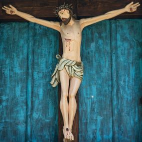 Zdjęcie nr 1: Krucyfiks drewniany w kapliczkowej oprawie umieszczony na absydzie, ponad sobotami. Chrystus przybity trzema gwoździami. Głowa opada na prawe ramię, oczy otwarte, nos długi, usta o kącikach skierowanych w dół. Twarz okala brązowy zarost. Na głowie ciemna korona cierniowa, włosy brązowe spływające na plecy i prawe ramię. Ciało umięśnione, nogi lekko ugięte. Z przebitego boku, spod korony oraz z miejsc przebicia gwoździami spływają strużki krwi. Perizonium białe, przewiązane sznurem, fałdujące się w równoległe pasy. Polichromia w partiach ciała naturalistyczna.