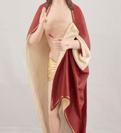 Zdjęcie nr 1: Rzeźba pełnoplastyczna przedstawiająca stojącego Chrystusa Zmartwychwstałego ustawiona na zielonej, owalnej podstawie i profilowanym, sześciobocznym cokoliku. Głowa zwrócona na wprost, twarz wydłużona, wzrok skierowany przed siebie, nos długi prosty, usta pełne. Twarz okala krótki brązowy zarost, włosy brązowe opadające na ramiona. Prawa ręka uniesiona w geście błogosławieństwa, lewa ugięta w łokciu na wysokość pasa, palce złączone. Na rękach, stopach i piersi widoczne ślady męczeństwa. Stopy nagie, biodra otacza złote, płasko sfałdowane perizonium. Płaszcz ciemnoczerwony ze złotą lamówką i białym podbiciem, rzeźbiony w głębokie fałdy. 