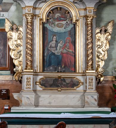 Zdjęcie nr 2: Obraz w kształcie stojącego prostokąta zamkniętego łukiem nadwieszonym, w złoconej i profilowanej ramie. W centrum kompozycji, u dołu przedstawienie św. Anny Samotrzeć. Maria z lewej i św. Anna z prawej siedzą obok siebie, na kolanach Marii stoi mały Jezus, który czyni gest błogosławieństwa w stronę św. Anny. Anna lewą ręką podaje mu owoc, w prawej trzyma otwartą książkę, patrzy na Jezusa. Maria jest ubrana w jasnofioletową suknię przewiązaną w pasie oraz ciemnobłękitny płaszcz z jasnym podbiciem, na głowie ma otwartą koronę. Święta Anna ma na sobie zielonkawą suknię oraz obszerny czerwony płaszcz narzucony na głowę, osłaniający ramiona oraz nogi. Jezus w białej, długiej, przylegającej do ciała tunice. Wszyscy mają nimby wokół głów. Pod ich stopami goła ziemia, na której widać kwitnące kwiaty. W górnej części kompozycji przedstawienie Boga Ojca i gołębicy Ducha Świętego w obłokach. Bóg jest widoczny do połowy ciała, ma rozłożone ręce i spogląda w dół, na siedzącą poniżej grupę. Ma długie siwe włosy oraz brodę, jest ubrany w niebieskawą tunikę i czerwony płaszcz, głowę rozświetla mu szeroki nimb, wokół którego unoszą się cztery uskrzydlone główki anielskie. Poniżej Boga Ojca gołębica z rozpostartymi skrzydłami w świetlistym otoku. Po ich bokach siedzą anioły grające na instrumentach.
 Kolorystyka ciemna, ciepła.