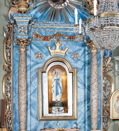 Zdjęcie nr 2: Ołtarz przyścienny, architektoniczny, jednoosiowy, jednokondygnacyjny ze zwieńczeniem i ażurowymi uszakami o falistym brzegu. Mensa prostopadłościenna z portatylem, antepedium ujęte profilowanymi listwami z polem wewnętrznym dekorowanym silnie stylizowanym, malowanym ornamentem o esowatym przebiegu i wolutowych końcach. Na mensie tabernakulum prostopadłościenne, na planie wklęsło-wypukłym, ujęte na narożach taśmami o skręconych końcach, zamknięte falującym gzymsem, pośrodku którego rocaille. Drzwiczki prostokątne, pozłacane z płaskorzeźbionym kielichem z hostią, zamknięte łukiem nadwieszonym. Tabernakulum marmoryzowane na jasnokremowy odcień. Retabulum ujęte parą kolumn kompozytowych ustawionych na niskich cokołach z rozetką, wspierających belkowanie wyłamujące się ponad nimi, z wydatnym, profilowanym gzymsem. Fryz dekorowany girlandami kwiatów w części środkowej, po bokach rozetkami. Nad belkowaniem na osiach kolumn ustawione wazony o żłobionej w dwóch strefach powierzchni. Pole środkowe z niszą ujętą prostokątną, profilowaną ramą, zamkniętą łukiem nadwieszonym. W niszy figura Matki Boskiej z Lourdes. Nad łukiem pozłacana korona otwarta i rozetki. Zwieńczenie półkoliste, ujęte spływami wolutowymi, na których rzeźby uskrzydlonych aniołków. Pośrodku zwieńczenia owalny obraz z przedstawieniem św. Józefa z Dzieciątkiem ujęty ramą uformowaną z obłoków z odchodzącymi promieniami. Nad zwieńczeniem rzeźbiona, złocona korona zamknięta. Całość struktury ołtarza marmoryzowana na kolor niebieski z ciemniejszymi żyłkami; kolumny jasnobrązowe z malowaną z wicią roślinną; ornamenty i profile złocone i srebrzone. 