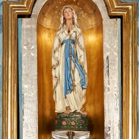 Zdjęcie nr 1: Rzeźba pełna na cokole przedstawiająca Matkę Boską  stojącą frontalnie, w kontrapoście, z lekko ugiętą lewą nogą i rękami złożonymi do modlitwy; przez lewą rękę ma przełożony różaniec. Maria ma lekko uniesioną i przechyloną głowę, oczy skierowane w górę, nos prosty, usta wąskie. Jest ubrana w białą suknię przewiązaną niebieskim pasem ze złotą lamówką i gwiazdkami, którego jeden, długi koniec opada w dół, a drugi nieco do boku. Ma również narzucony na głowę oraz ramiona, opadający miękkimi fałdami biały płaszcz dekorowany na brzegach pasem złotego ornamentu. Spod sukni wystają bose stopy, na każdej z nich złoty kwiat róży. Mara stoi na cokole imitującym kamień położonym na prostopadłościanie o ściętych narożach zdobionym różami na zielonym tle. Polichromia w partiach ciała naturalistyczna.