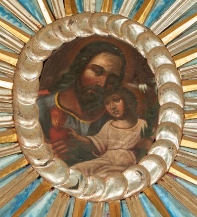 Zdjęcie nr 1: Obraz w kształcie owalu przedstawiający św. Józefa z Dzieciątkiem w ramie utworzonej ze srebrnych obłoków. Święty został  ukazanego w popiersiu, frontalnie, z głową przechyloną w lewo, dotykającą głowy Dzieciątka. Jest ubrany w niebieską tunikę lamowaną złotem pod szyją, za jego plecami widać fragment czerwonego płaszcza. Dzieciątko siedzi zwrócone w trzech czwartych w prawo, ukazane do kolan,  z prawą ręką wyciągniętą w kierunku trzymanego przez Józefa gorejącego serca i lilią w lewej. Jest ubrane w białą, przepasaną tunikę z szerokimi rękawami; od głowy Dzieciątka odchodzą promienie. Tło obrazu ciemnobrązowe, rozjaśnione wokół głów postaci. 