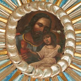 Zdjęcie nr 1: Obraz w kształcie owalu przedstawiający św. Józefa z Dzieciątkiem w ramie utworzonej ze srebrnych obłoków. Święty został  ukazanego w popiersiu, frontalnie, z głową przechyloną w lewo, dotykającą głowy Dzieciątka. Jest ubrany w niebieską tunikę lamowaną złotem pod szyją, za jego plecami widać fragment czerwonego płaszcza. Dzieciątko siedzi zwrócone w trzech czwartych w prawo, ukazane do kolan,  z prawą ręką wyciągniętą w kierunku trzymanego przez Józefa gorejącego serca i lilią w lewej. Jest ubrane w białą, przepasaną tunikę z szerokimi rękawami; od głowy Dzieciątka odchodzą promienie. Tło obrazu ciemnobrązowe, rozjaśnione wokół głów postaci. 