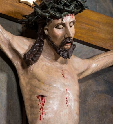 Zdjęcie nr 3: Rzeźba pełnoplastyczna, ścięta w partii pleców, polichromowana figura Chrystusa na prostym krzyżu, przybita do niego trzema metalowymi gwoździami. Sylwetka zmarłego Jezusa jest smukła, wychudzona, z wyraźnie zarysowaną klatką piersiową. Ramiona rozpostarte, uniesione ponad głowę. Nogi z zaznaczonym zarysem mięśni są lekko ugięte w kolanach. Głowa opadająca, przechylona w prawą stronę, z szeroką koroną cierniową o gęstym splocie. Twarz pociągła z krótkim zarostem, oczy zamknięte, nos spiczasty o prostym profilu, usta otwarte, o opadłych kącikach. Włosy długie, rzeźbione w formie sznurów opadają na plecy i prawe ramię. Perizonium krótkie, okrywające górną część nóg, udrapowane na pojedynczym sznurze, fałdy płasko modelowane, zachodzące na siebie. Nad głową Zbawiciela pozłacany titulus z napisem „INRI”. Figura zmarłego Chrystusa pokryta polichromią imitującą naturalną karnację, ciemnoczerwoną farbą zaznaczone ślady męki na dłoniach, stopach, prawym boku i głowie. Perizonium pozłacane, włosy i zarost ciemnobrązowe, korona cierniowa czarna. Krucyfiks umieszczono na tle obrazu z przedstawieniem stojących pod krzyżem postaci Marii po lewej i św. Jana po prawej stronie. Maria stoi wyprostowana, spogląda na Jezusa, ręce składa na piersi. Ma pogodną, owalną twarz, duże oczy i prosty nos. Jest ubrana w różową suknię i bardzo obszerny niebieski płaszcz, który okrywa jej lewe ramię, plecy i prawą nogę, łamiąc się w grube fałdy. Na głowie ma biały welon, spod sukni wystają stopy w sandałach. Święty Jan stoi w wykroku, z prawą nogą z przodu. Lewą rękę ma odwiedzioną do tyłu, prawą przykłada do twarzy fragment płaszcza. Ma małą głowę, jasne długie włosy opadają mu na plecy. Jest ubrany w zieloną, przewiązaną w pasie suknię i czerwony płaszcz przytroczony przy lewym biodrze. Postacie stoją na tle widocznej w oddali panoramy miasta, reszta tła brunatna. 