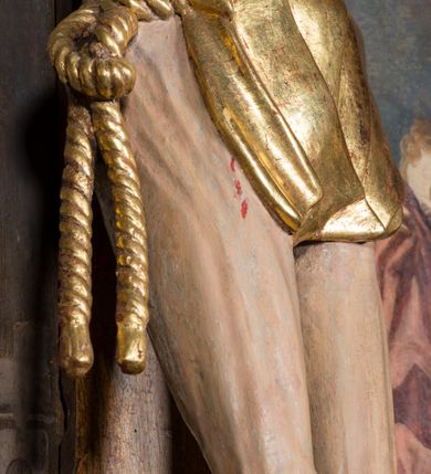 Zdjęcie nr 4: Rzeźba pełnoplastyczna, ścięta w partii pleców, polichromowana figura Chrystusa na prostym krzyżu, przybita do niego trzema metalowymi gwoździami. Sylwetka zmarłego Jezusa jest smukła, wychudzona, z wyraźnie zarysowaną klatką piersiową. Ramiona rozpostarte, uniesione ponad głowę. Nogi z zaznaczonym zarysem mięśni są lekko ugięte w kolanach. Głowa opadająca, przechylona w prawą stronę, z szeroką koroną cierniową o gęstym splocie. Twarz pociągła z krótkim zarostem, oczy zamknięte, nos spiczasty o prostym profilu, usta otwarte, o opadłych kącikach. Włosy długie, rzeźbione w formie sznurów opadają na plecy i prawe ramię. Perizonium krótkie, okrywające górną część nóg, udrapowane na pojedynczym sznurze, fałdy płasko modelowane, zachodzące na siebie. Nad głową Zbawiciela pozłacany titulus z napisem „INRI”. Figura zmarłego Chrystusa pokryta polichromią imitującą naturalną karnację, ciemnoczerwoną farbą zaznaczone ślady męki na dłoniach, stopach, prawym boku i głowie. Perizonium pozłacane, włosy i zarost ciemnobrązowe, korona cierniowa czarna. Krucyfiks umieszczono na tle obrazu z przedstawieniem stojących pod krzyżem postaci Marii po lewej i św. Jana po prawej stronie. Maria stoi wyprostowana, spogląda na Jezusa, ręce składa na piersi. Ma pogodną, owalną twarz, duże oczy i prosty nos. Jest ubrana w różową suknię i bardzo obszerny niebieski płaszcz, który okrywa jej lewe ramię, plecy i prawą nogę, łamiąc się w grube fałdy. Na głowie ma biały welon, spod sukni wystają stopy w sandałach. Święty Jan stoi w wykroku, z prawą nogą z przodu. Lewą rękę ma odwiedzioną do tyłu, prawą przykłada do twarzy fragment płaszcza. Ma małą głowę, jasne długie włosy opadają mu na plecy. Jest ubrany w zieloną, przewiązaną w pasie suknię i czerwony płaszcz przytroczony przy lewym biodrze. Postacie stoją na tle widocznej w oddali panoramy miasta, reszta tła brunatna. 