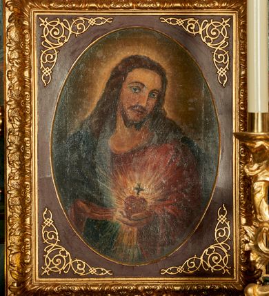 Zdjęcie nr 2: Obraz z przedstawieniem Najświętszego Serca Jezusa ukazanego w półpostaci, frontalnie z głową przechyloną w stronę lewego barku, sercem w lewej dłoni i wskazujący na nie prawą dłonią. Głowa zwrócona delikatnie w lewo, wzrok skierowany na widza, nos prosty, usta wąskie. Twarz okala bujny, ciemnobrązowy zarost, falowane włosy opadają na ramiona i plecy. Wokół głowy znajduje się świetlisty nimb. Chrystus ubrany jest w czerwoną suknię z długimi rękawami i granatowy płaszcz, który przerzucony ma przez prawy bark. Trzymane w dłoni serce jest otoczone koroną cierniową i zwieńczone krzyżem łacińskim, otacza je jasna, promienista gloria. Tło ciemnobrązowe, jaśniejące wokół głowy Chrystusa. Obraz ujęty jest w bogato zdobione prostokątne i profilowane ramy o owalnym świetle, dekorowane w narożach stylizowanym ornamentem cęgowym.