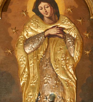 Zdjęcie nr 4: Obraz w kształcie stojącego prostokąta zamkniętego łukiem nadwieszonym w profilowanej, złoconej ramie. W centrum kompozycji ukazana Matka Boska w całej postaci, stojąca na półksiężycu z zaznaczonymi rysami twarzy oraz globie, depcząca węża trzymającego w paszczy jabłko. Obie ręce ma złożone na piersi, głowę skłania. Ma owalną, delikatną twarz, spogląda w dół, ma ciemne oczy przesłonięte dużymi powiekami, wąski nos i niewielkie usta oraz długie, ciemne włosy opadające na plecy. Jej głowę otacza świetlisty nimb, ramiona i głowę wieniec z 10 gwiazd. Ubranie jest przysłonięte płaskorzeźbioną, drewnianą sukienkę w postaci tuniki i płaszcza dekorowanych kwiatami, stopy bose. Tło ciemnobrązowe, niejednolite, kolorystyka ciepła, brunatna.