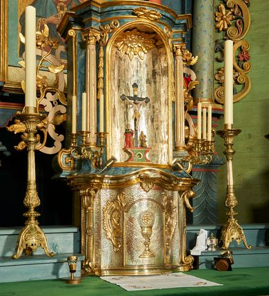Zdjęcie nr 2: Tabernakulum o rozbudowanej, dwustrefowej strukturze, na planie wklęsło-wypukłym. Dolna kondygnacja niższa, z wypukłymi drzwiczkami pośrodku, górna z ulokowanym nad nimi tronem wystawienia w formie wnęki zamkniętej łukiem półkolistym. Każdą kondygnację wieńczy pełne belkowanie, wyłamane na osiach ułożonych skośnie wolut w dolnej partii i kolumienek korynckich w górnej. Na drzwiczkach tabernakulum znajduje się dekoracja złożona z kielicha z hostią otoczoną glorią promienistą, w niej monogram „IHS”. Kielich stoi na zwężającej się ku górze stopie, trzon w połowie wysokości przerywa nodus, a czarę ujmuje koszyczek. Drzwiczki flankują plakiety ornamentalne skomponowane z płomienistego rocaille’a. Wynoszące się na osi belkowanie spina stylizowany liść akantu. W niszę powyżej wstawiono rzeźbę przedstawiającą grupę ukrzyżowania, sklepienie dekoruje plakieta z liści i kwiatów. Niszę otaczają fragmenty wici, z której zwieszają się rzędy kampanul. Na belkowaniu tej kondygnacji plakieta z liści. kondygnacja ujęta spływami wolutowymi zakończonymi meandrem, tron uszakami w postaci cęgów przeplatanych gałązek różanych. Dolna część srebrzona i złocona; struktura górnej marmoryzowana na kolor granatowoniebieski, kolumny szare, profile i ornamenty złocone. 