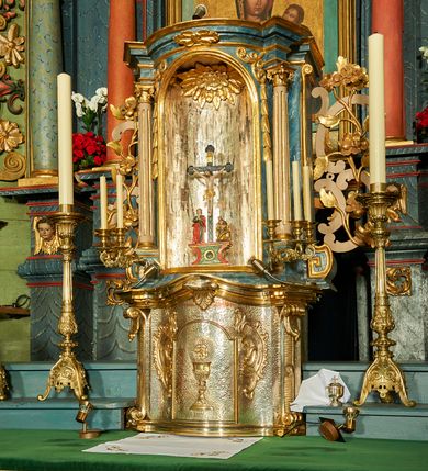 Zdjęcie nr 1: Tabernakulum o rozbudowanej, dwustrefowej strukturze, na planie wklęsło-wypukłym. Dolna kondygnacja niższa, z wypukłymi drzwiczkami pośrodku, górna z ulokowanym nad nimi tronem wystawienia w formie wnęki zamkniętej łukiem półkolistym. Każdą kondygnację wieńczy pełne belkowanie, wyłamane na osiach ułożonych skośnie wolut w dolnej partii i kolumienek korynckich w górnej. Na drzwiczkach tabernakulum znajduje się dekoracja złożona z kielicha z hostią otoczoną glorią promienistą, w niej monogram „IHS”. Kielich stoi na zwężającej się ku górze stopie, trzon w połowie wysokości przerywa nodus, a czarę ujmuje koszyczek. Drzwiczki flankują plakiety ornamentalne skomponowane z płomienistego rocaille’a. Wynoszące się na osi belkowanie spina stylizowany liść akantu. W niszę powyżej wstawiono rzeźbę przedstawiającą grupę ukrzyżowania, sklepienie dekoruje plakieta z liści i kwiatów. Niszę otaczają fragmenty wici, z której zwieszają się rzędy kampanul. Na belkowaniu tej kondygnacji plakieta z liści. kondygnacja ujęta spływami wolutowymi zakończonymi meandrem, tron uszakami w postaci cęgów przeplatanych gałązek różanych. Dolna część srebrzona i złocona; struktura górnej marmoryzowana na kolor granatowoniebieski, kolumny szare, profile i ornamenty złocone. 