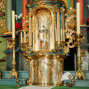 Zdjęcie nr 1: Tabernakulum o rozbudowanej, dwustrefowej strukturze, na planie wklęsło-wypukłym. Dolna kondygnacja niższa, z wypukłymi drzwiczkami pośrodku, górna z ulokowanym nad nimi tronem wystawienia w formie wnęki zamkniętej łukiem półkolistym. Każdą kondygnację wieńczy pełne belkowanie, wyłamane na osiach ułożonych skośnie wolut w dolnej partii i kolumienek korynckich w górnej. Na drzwiczkach tabernakulum znajduje się dekoracja złożona z kielicha z hostią otoczoną glorią promienistą, w niej monogram „IHS”. Kielich stoi na zwężającej się ku górze stopie, trzon w połowie wysokości przerywa nodus, a czarę ujmuje koszyczek. Drzwiczki flankują plakiety ornamentalne skomponowane z płomienistego rocaille’a. Wynoszące się na osi belkowanie spina stylizowany liść akantu. W niszę powyżej wstawiono rzeźbę przedstawiającą grupę ukrzyżowania, sklepienie dekoruje plakieta z liści i kwiatów. Niszę otaczają fragmenty wici, z której zwieszają się rzędy kampanul. Na belkowaniu tej kondygnacji plakieta z liści. kondygnacja ujęta spływami wolutowymi zakończonymi meandrem, tron uszakami w postaci cęgów przeplatanych gałązek różanych. Dolna część srebrzona i złocona; struktura górnej marmoryzowana na kolor granatowoniebieski, kolumny szare, profile i ornamenty złocone. 