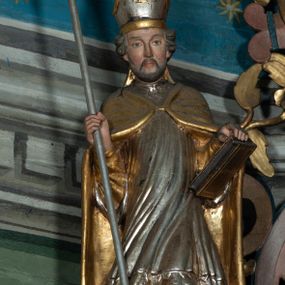 Zdjęcie nr 1: Rzeźba pełna, święty zwrócony na wprost, w kontrapoście. Lewą ręką podtrzymuje księgę opartą na biodrze, w prawej trzyma krzyż arcybiskupi. Twarz pociągła, o wyraźnych rysach, okolona zarostem; włosy krótkie, falowane. Święty jest ubrany w złotą albę, srebrną komżę dekorowaną u spodu ornamentem roślinnym oraz złotą kapę zapiętą na piersi fibulą; na głowie ma infułę. Polichromia naturalistyczna w partiach ciała.