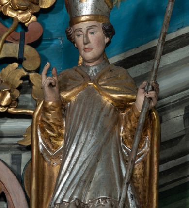 Zdjęcie nr 1: Rzeźba całopostaciowa, zwrócona na wprost, w kontrapoście. Prawa ręka zgięta w geście błogosławieństwa, lewa przytrzymuje pastorał zakończony krzywaśnią w formie liścia akantu z małżowiną. Twarz pociągła, o na wpół przymkniętych oczach; włosy krótkie. Święty jest ubrany w złotą albę, srebrną komżę, zdobioną u spodu koronką oraz przykryty złotą kapą spiętą fibulą; na głowie ma infułę. Polichromia naturalistyczna w partiach ciała. 