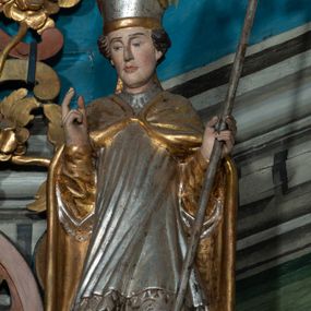 Zdjęcie nr 1: Rzeźba całopostaciowa, zwrócona na wprost, w kontrapoście. Prawa ręka zgięta w geście błogosławieństwa, lewa przytrzymuje pastorał zakończony krzywaśnią w formie liścia akantu z małżowiną. Twarz pociągła, o na wpół przymkniętych oczach; włosy krótkie. Święty jest ubrany w złotą albę, srebrną komżę, zdobioną u spodu koronką oraz przykryty złotą kapą spiętą fibulą; na głowie ma infułę. Polichromia naturalistyczna w partiach ciała. 