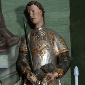 Zdjęcie nr 1: Rzeźba drążona, ścięta z tyłu. Figura zwrócona na wprost, z głową skręconą lekko w prawo. Ręce opuszczone na dół, w prawej zielona palma męczeństwa, w lewej szare kamienie. Twarz pełna, o małych oczach, długim nosie i wąskich ustach. Włosy krótkie, kręcone. Święty jest ubrany w szarą albę, srebrzoną u dołu z rękawami zakończonymi złotą lamówką, srebrny humerał oraz dalmatykę dekorowaną ukwieconymi gałązkami. Polichromia naturalistyczna w partiach ciała.