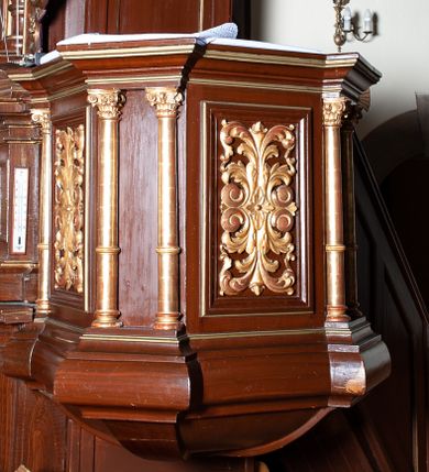 Zdjęcie nr 1: Ambona zawieszona w arkadzie tęczy, dostępna jednobiegowymi schodami od wschodu, od strony wejścia do zakrystii. Kosz na rzucie ośmioboku, opięty smukłymi kolumienkami o gładkich trzonach i kompozytowych kapitelach, ustawionymi występujących wybrzuszonych postumentach i wspierającymi gzyms występujący na nimi. Boki kosza na przemian gładkie i ozdobione nałożonymi rzeźbionymi motywami roślinnymi. Kosz nadwieszony na konsoli w kształcie spłaszczonego ostrosłupa. Zaplecek prostokątny, z prostokątną zamkniętą półkoliście płyciną, w której w górnej części litery IHS z krzyżem, otoczony promieniami. Baldachim powtarza rzut i podziały kosza, ozdobiomy lambrekinem, nakryty spłaszczonym ostrosłupem zwieńczonym prostym krzyżykiem. Na podniebiu rzeźbiona Gołębica Ducha Świętego wśród promieni. Struktura pomalowana na brązowo, podniebie baldachimu w kolorze bękitnym, ornamenty i detale architektoniczne złocone. 