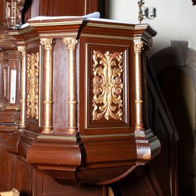 Zdjęcie nr 1: Ambona zawieszona w arkadzie tęczy, dostępna jednobiegowymi schodami od wschodu, od strony wejścia do zakrystii. Kosz na rzucie ośmioboku, opięty smukłymi kolumienkami o gładkich trzonach i kompozytowych kapitelach, ustawionymi występujących wybrzuszonych postumentach i wspierającymi gzyms występujący na nimi. Boki kosza na przemian gładkie i ozdobione nałożonymi rzeźbionymi motywami roślinnymi. Kosz nadwieszony na konsoli w kształcie spłaszczonego ostrosłupa. Zaplecek prostokątny, z prostokątną zamkniętą półkoliście płyciną, w której w górnej części litery IHS z krzyżem, otoczony promieniami. Baldachim powtarza rzut i podziały kosza, ozdobiomy lambrekinem, nakryty spłaszczonym ostrosłupem zwieńczonym prostym krzyżykiem. Na podniebiu rzeźbiona Gołębica Ducha Świętego wśród promieni. Struktura pomalowana na brązowo, podniebie baldachimu w kolorze bękitnym, ornamenty i detale architektoniczne złocone. 