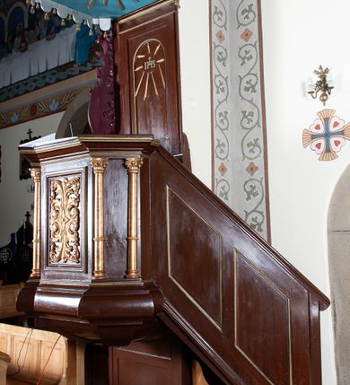 Zdjęcie nr 2: Ambona zawieszona w arkadzie tęczy, dostępna jednobiegowymi schodami od wschodu, od strony wejścia do zakrystii. Kosz na rzucie ośmioboku, opięty smukłymi kolumienkami o gładkich trzonach i kompozytowych kapitelach, ustawionymi występujących wybrzuszonych postumentach i wspierającymi gzyms występujący na nimi. Boki kosza na przemian gładkie i ozdobione nałożonymi rzeźbionymi motywami roślinnymi. Kosz nadwieszony na konsoli w kształcie spłaszczonego ostrosłupa. Zaplecek prostokątny, z prostokątną zamkniętą półkoliście płyciną, w której w górnej części litery IHS z krzyżem, otoczony promieniami. Baldachim powtarza rzut i podziały kosza, ozdobiomy lambrekinem, nakryty spłaszczonym ostrosłupem zwieńczonym prostym krzyżykiem. Na podniebiu rzeźbiona Gołębica Ducha Świętego wśród promieni. Struktura pomalowana na brązowo, podniebie baldachimu w kolorze bękitnym, ornamenty i detale architektoniczne złocone. 