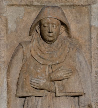 Zdjęcie nr 1: Płyta nagrobna w kształcie stojącego prostokąta, z listwowym obramieniem, na którym częściowo zatarta łacińska inskrypcja majuskułowa: ANNO DOMINI…. W polu płaskorzeźbione przedstawienie kobiety w całej postaci, ubranej w narzuconą na ramiona, rozciętą z przodu jubkę-kaftanik oraz w obszerną długą spódnicę z fałdami na bokach. Na głowie czepiec z podwiką.  Twarz dojrzałej kobiety, pełna, z wydatnymi uszami. Spod rozcięcia wysunięte i złożone na piersiach dłonie w rękawiczkach.