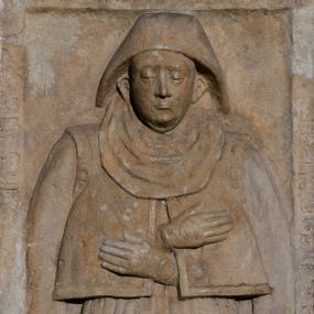 Zdjęcie nr 1: Płyta nagrobna w kształcie stojącego prostokąta, z listwowym obramieniem, na którym częściowo zatarta łacińska inskrypcja majuskułowa: ANNO DOMINI…. W polu płaskorzeźbione przedstawienie kobiety w całej postaci, ubranej w narzuconą na ramiona, rozciętą z przodu jubkę-kaftanik oraz w obszerną długą spódnicę z fałdami na bokach. Na głowie czepiec z podwiką.  Twarz dojrzałej kobiety, pełna, z wydatnymi uszami. Spod rozcięcia wysunięte i złożone na piersiach dłonie w rękawiczkach.